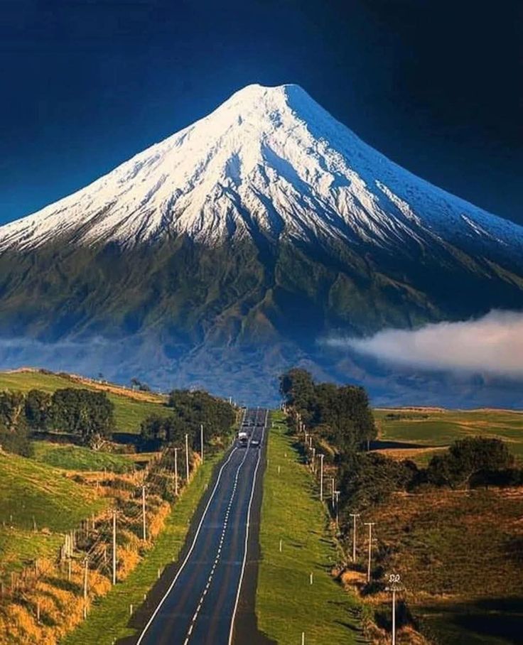 Straight road leading toward a snow-capped volcanic mountain with green hills and clouds