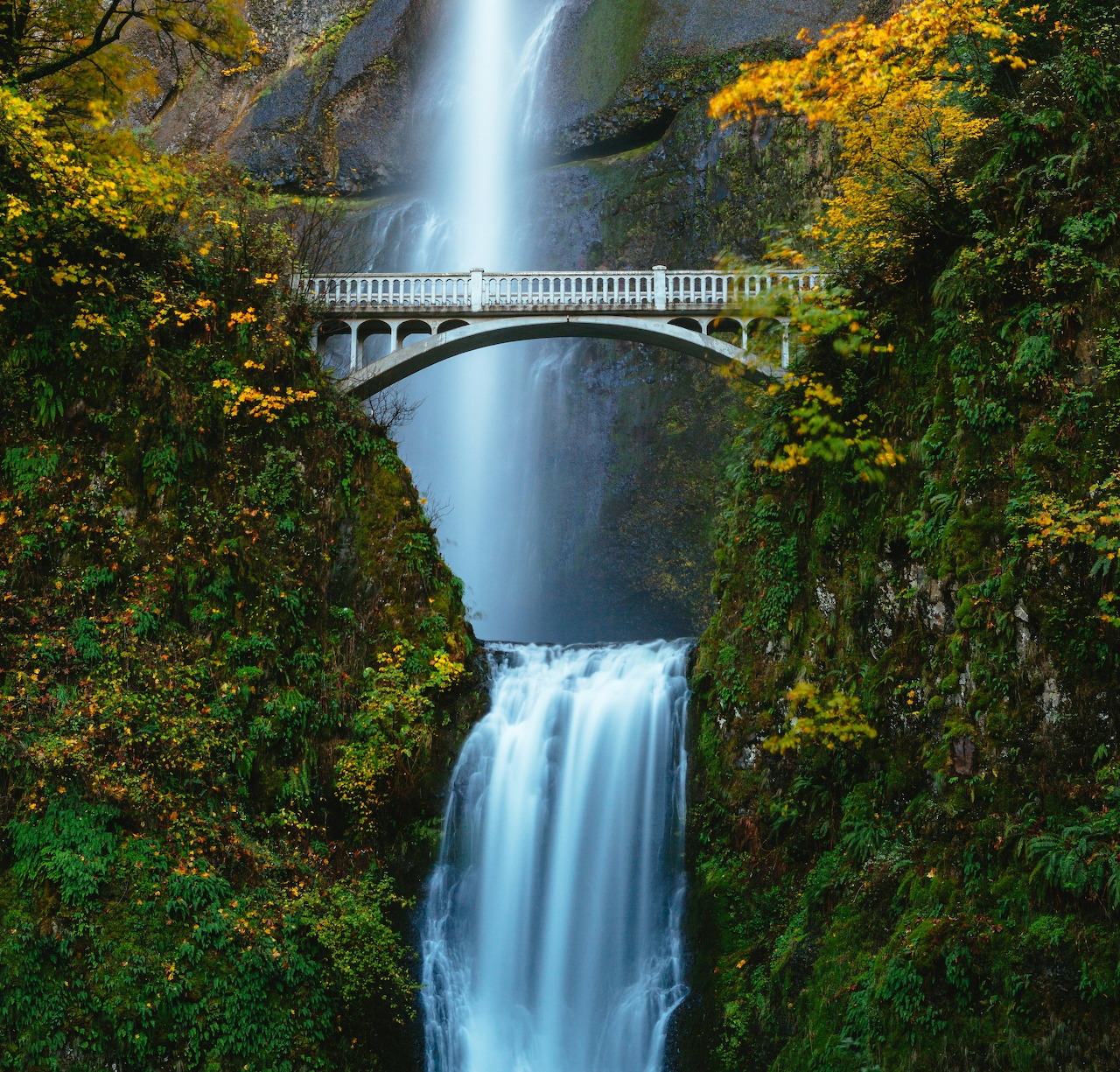 Two-tier waterfall flows under stone bridge surrounded by autumn leaves