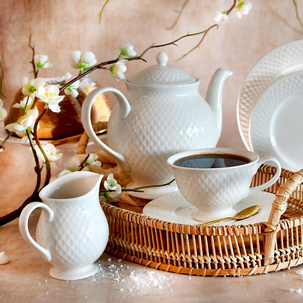 White textured teapot, creamer, and cup of black coffee on a wicker tray with cherry blossoms