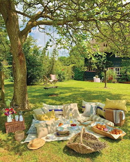 Picnic blanket with food and basket under tree on sunny lawn, cottage visible in background