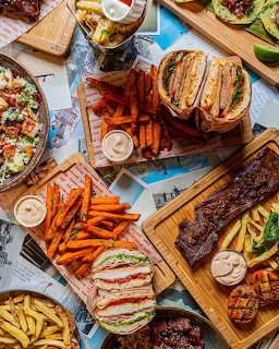 Overhead flat lay of grilled burgers, ribs, fries, pizza, and sandwiches on wooden serving boards