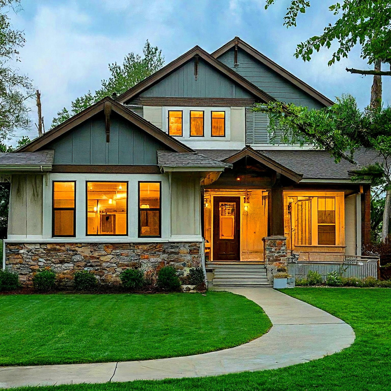 Craftsman house with stone facade and glowing windows at dusk, surrounded by green lawn and trees