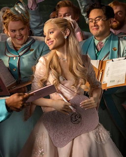 Blonde woman in a pale pink gown holding books, surrounded by people in teal uniforms indoors.
