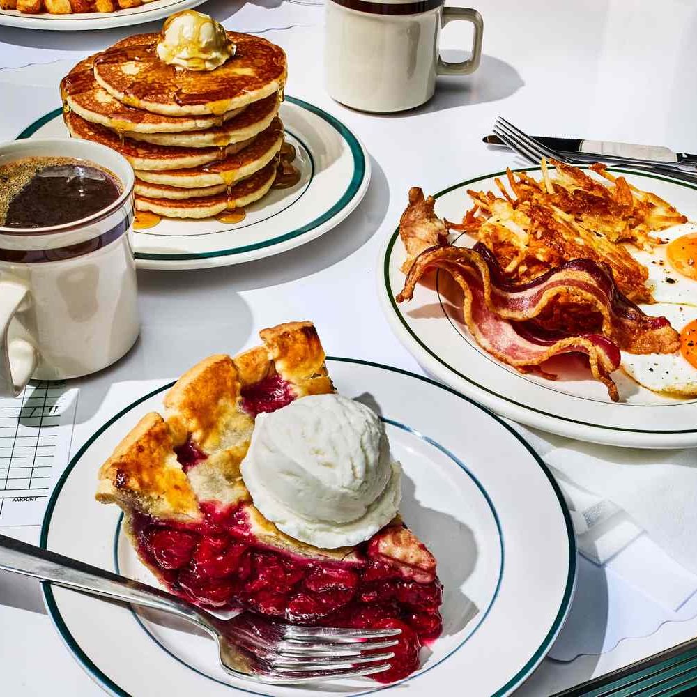 Breakfast table with pancakes, bacon eggs, berry pie, coffee and juice on white plates