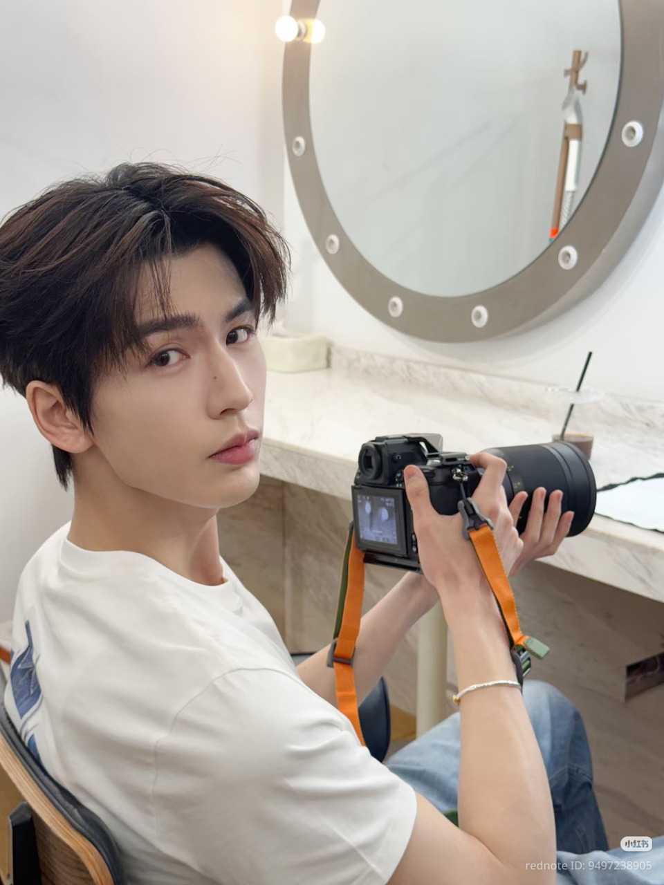 Young Asian man holding a DSLR camera on a tripod indoors, round mirror visible on the wall behind him.