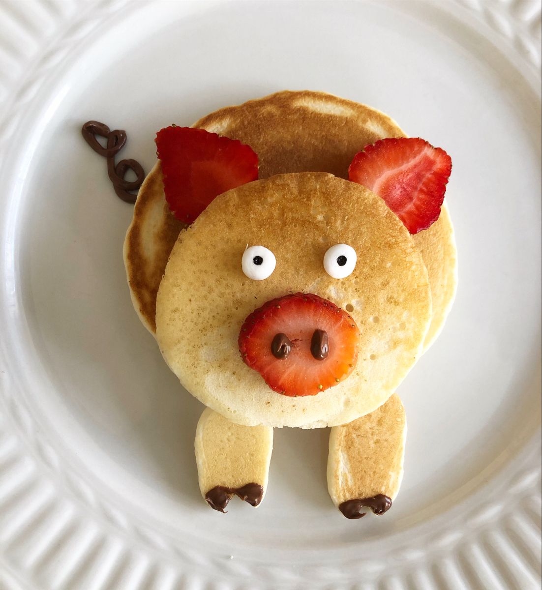 Pig-shaped pancakes on a white plate with strawberry ears, candy eyes, and a fruit snout