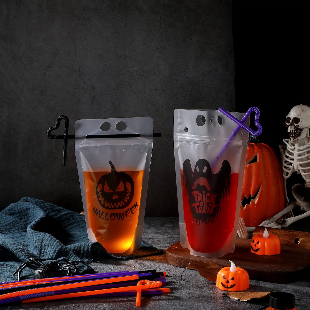 Two Halloween drinks in clear pouches with pumpkins, a skeleton, and striped straws on a dark table