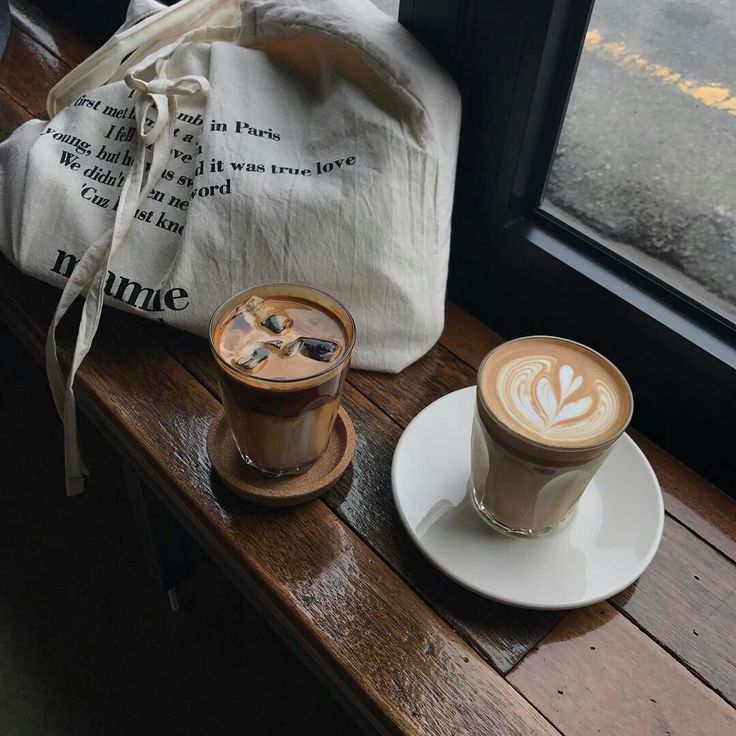 Two coffee drinks with latte art on wooden cafe table near window with tote bag