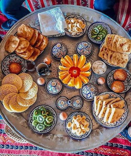Overhead view of a round tray with pastries, bread, fruit, tea, and patterned bowls on a colorful cloth