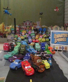 Colorful toy cars gathered on a play mat with a Radiator Springs sign and miniature buildings