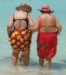 Two elderly women in colorful swimwear and straw hats wading in shallow clear turquoise water at a beach