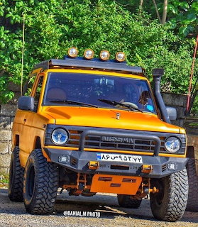 Modified yellow Nissan Patrol off-road vehicle with roof lights parked in front of green trees