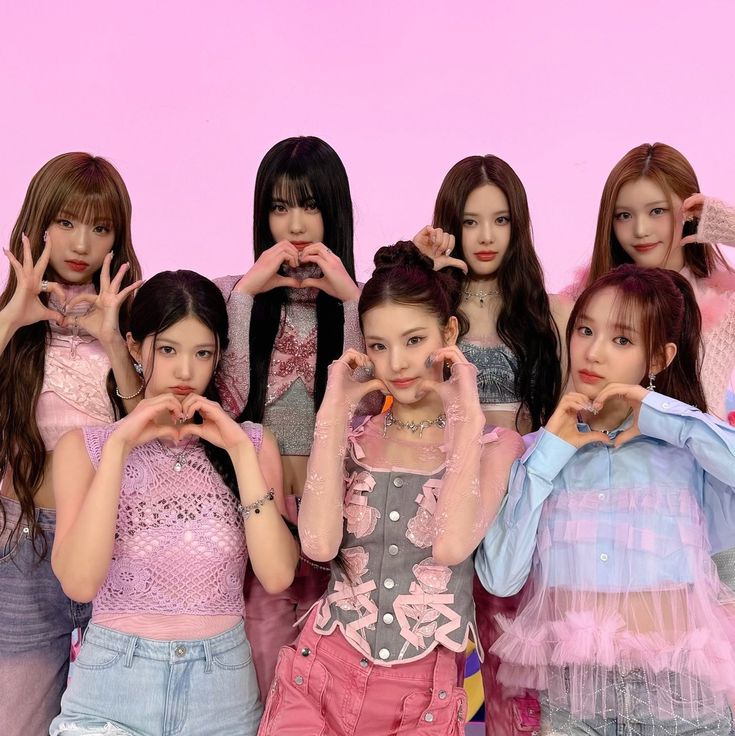 Six young women in pastel outfits posing with heart hand gestures against a pink background