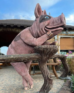 Large pink pig statue leaning casually on a wooden fence outside a rustic building