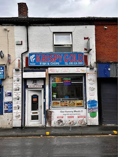 Small fish and chips shopfront with signs, window posters, and neighboring brick buildings
