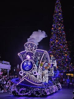 Glowing parade float resembling a train next to a lit Christmas tree at night in a Disney park