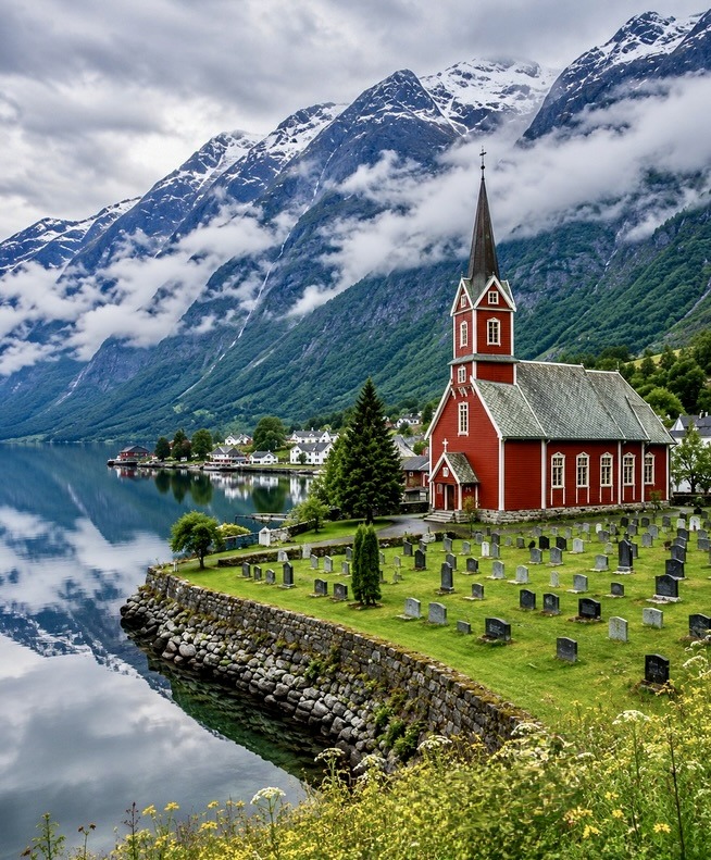 Red wooden church with steeple by a Norwegian fjord surrounded by snow-capped mountains and a cemetery