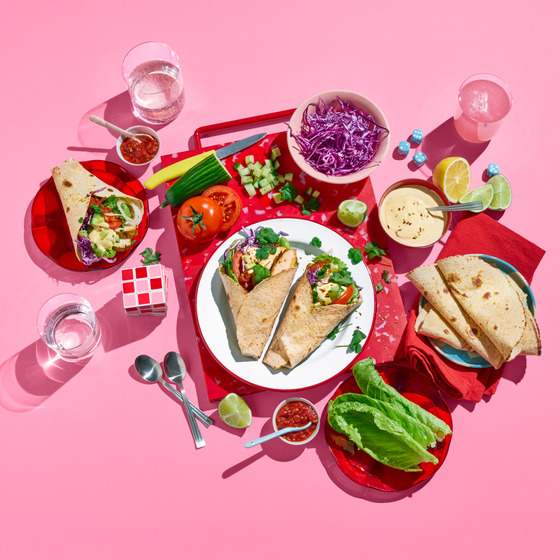 Overhead shot of chicken wraps with vegetables, dips, and flatbread on red plates on pink background