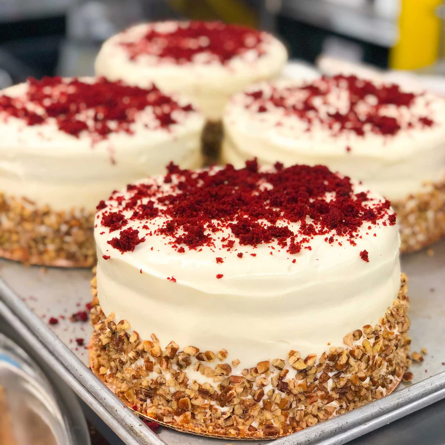 Four round red velvet cakes with cream cheese frosting, red crumbs on top, and pecan-coated sides on a serving tray