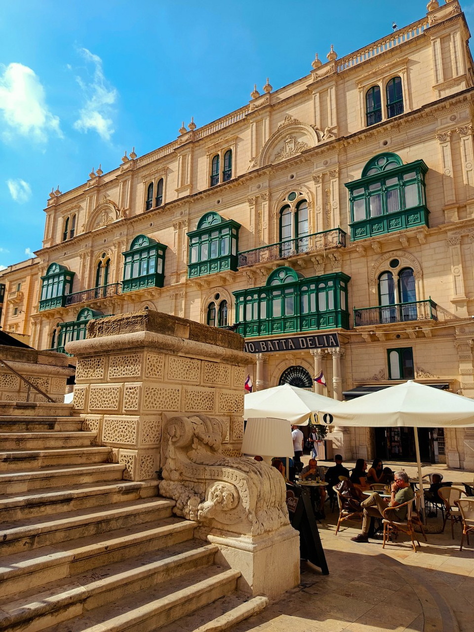 Historic Valletta building with stone stairs, green balconies, and cafe umbrellas under a blue sky