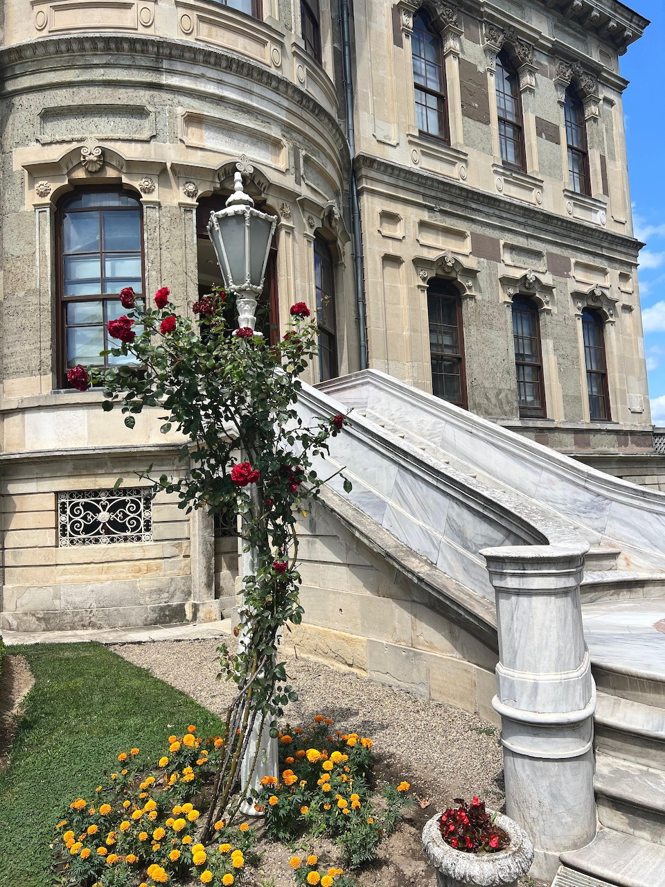 Ornate stone facade of Küçüksu Palace with marble staircase, lamp post with climbing roses, and garden flowers