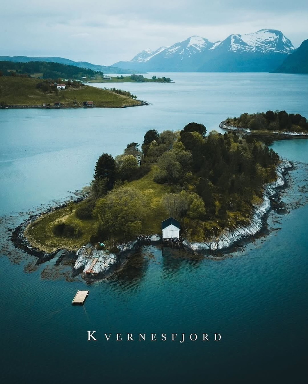 Aerial view of a small island with trees and a white boathouse on Kvernesfjord, Norway, with snowy mountains behind