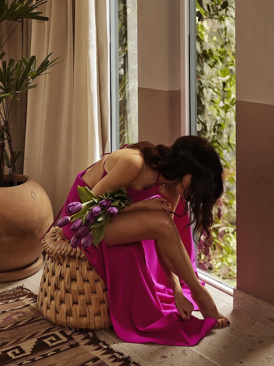 Woman in a bright pink dress sitting by an open doorway, holding purple flowers
