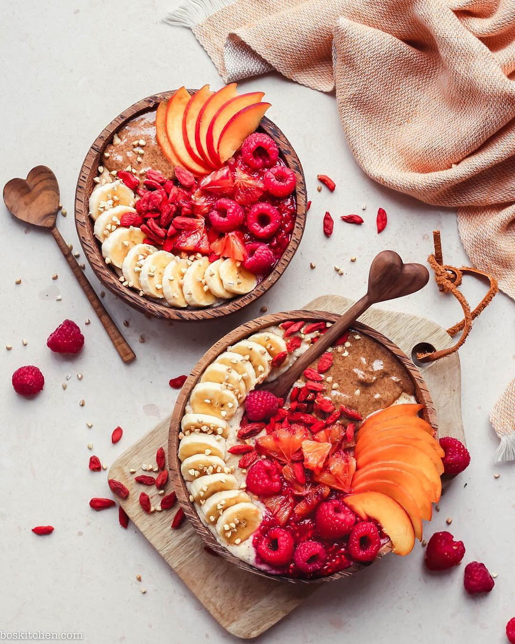 Two smoothie bowls with fresh fruit arranged in colorful patterns including bananas, persimmons, and raspberries