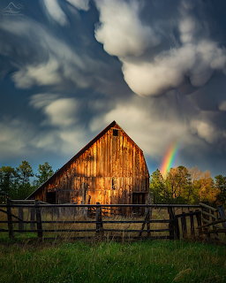 Old wooden barn with rainbow appearing in stormy evening sky, wooden fence in foreground