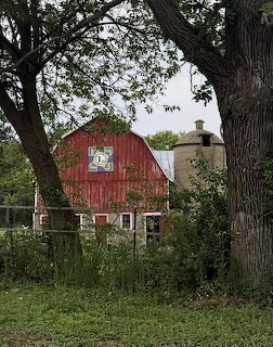 Red barn with painted quilt square on front, framed by trees, with a silo beside it on a cloudy day