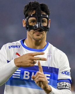 Soccer player in white jersey with blue stripes and protective face mask making hand gesture
