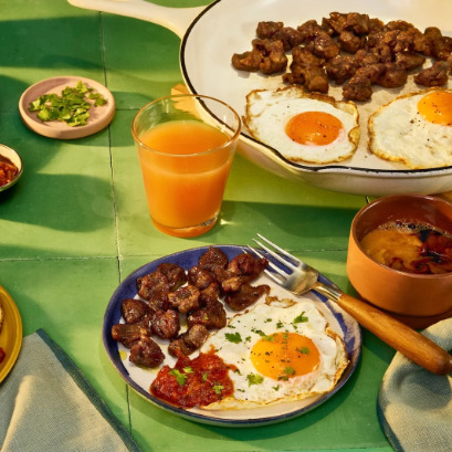 Breakfast plate with fried eggs and sausage alongside orange juice, beans, and garnishes on a green table