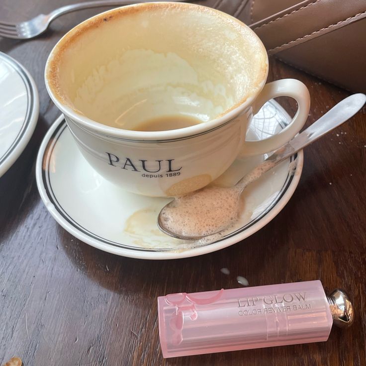 White coffee cup labeled Paul on saucer with pink lipgloss tube and spoon on wooden table