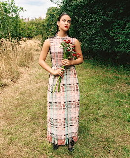 Woman in pink plaid dress holding flowers standing in garden with trees and grass in background