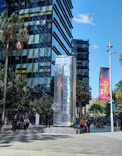 Transparent bus-shaped sculpture in an urban plaza with glass buildings, palm trees, and blue sky