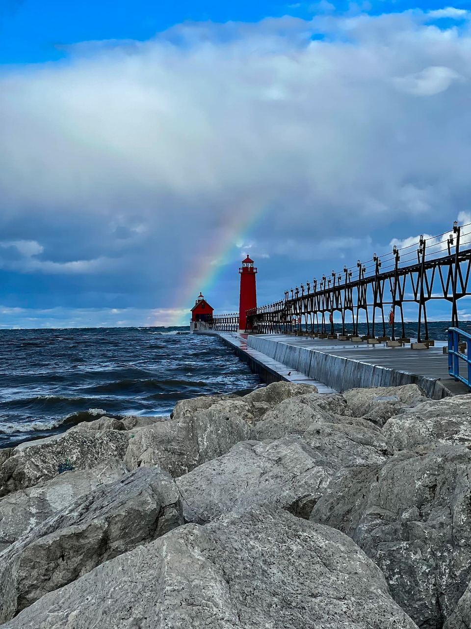 Red lighthouse on pier over ocean with rocks in foreground and cloudy sky