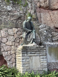 Seated statue on a stone pedestal against a rock wall beside a small waterfall