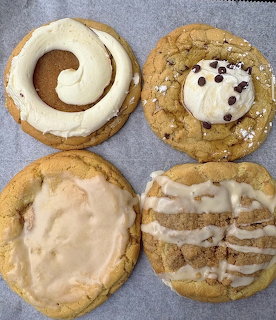 Four large frosted gourmet cookies on parchment paper with various toppings including cream frosting and chocolate chips