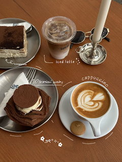 Overhead café table with tiramisu slices, iced latte, cappuccino with latte art, and a candle on grey plates.