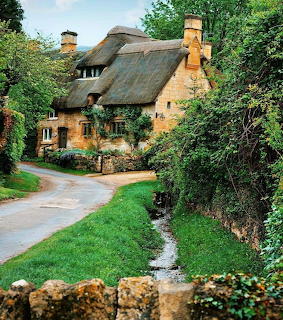 Quaint thatched cottage with stone walls along a winding road surrounded by green foliage