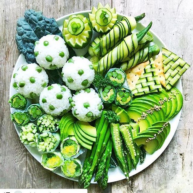 Circular plate of assorted green vegetables and fruits artfully arranged on white rustic wood surface