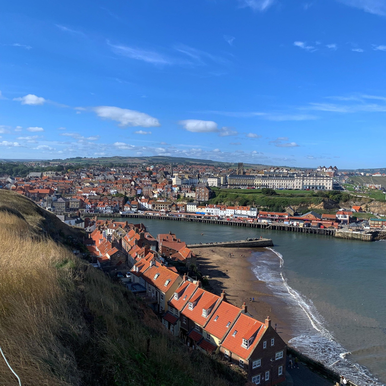 Hilltop view of a coastal town with red roofs, shoreline, river, and blue sky