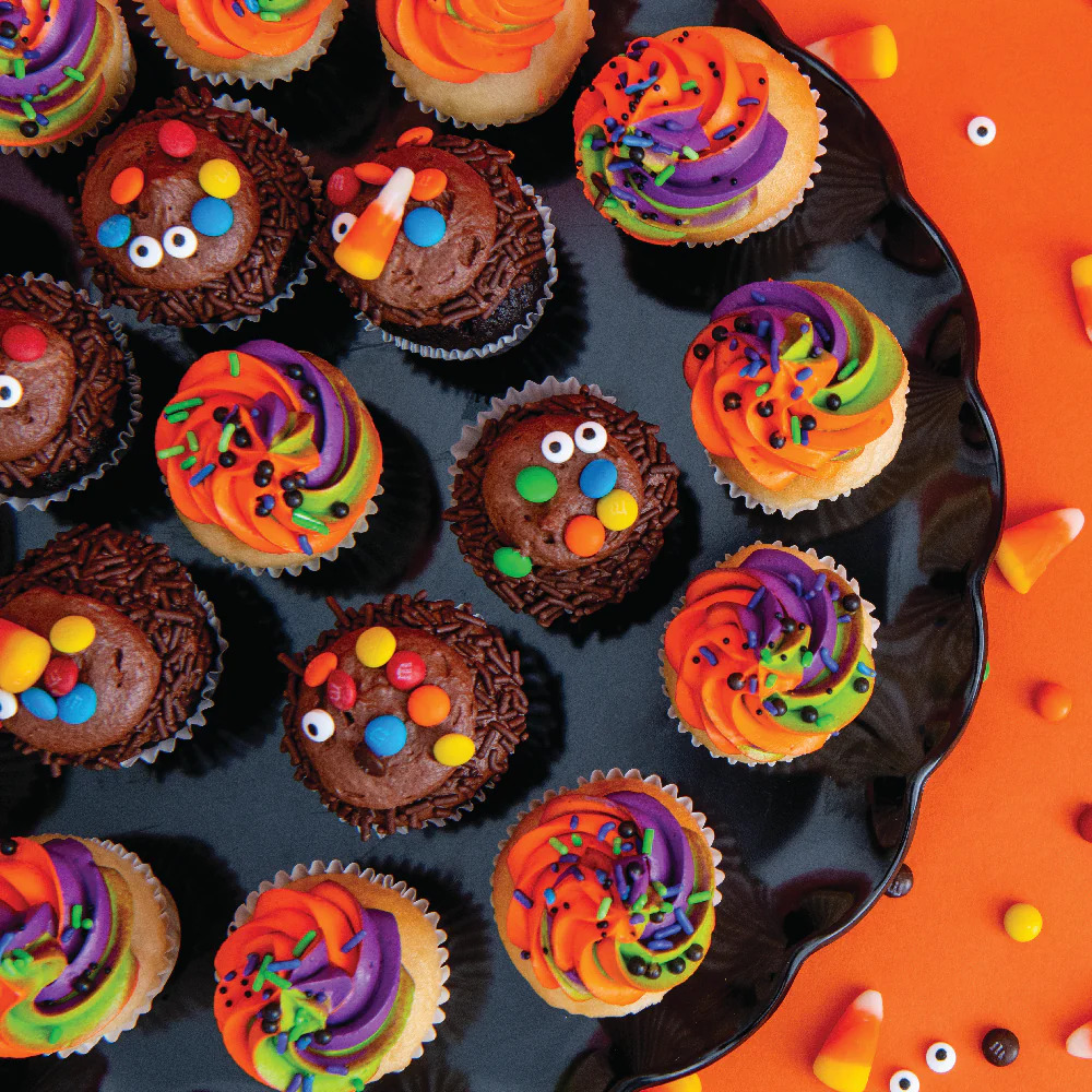 Halloween cupcakes with rainbow frosting, candy eyes, and colorful sprinkles arranged on black surface