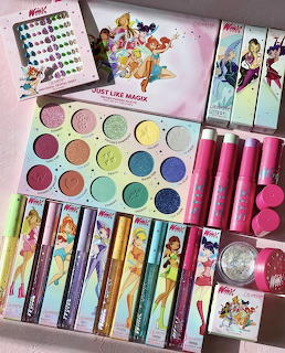 Pastel Winx-themed makeup collection with eyeshadow palette, lipsticks and mascaras on pink background