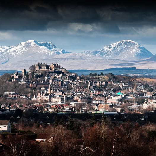 Stirling Castle on hilltop with Scottish town below and snowy mountains under dark storm clouds