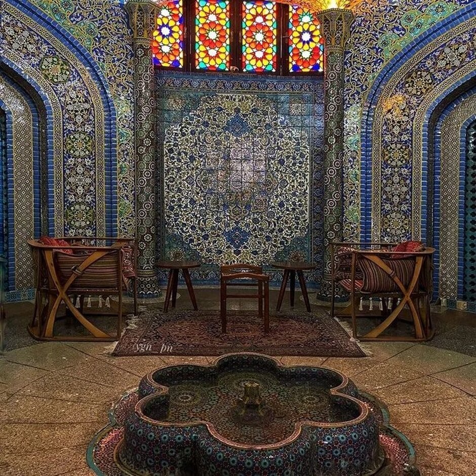 Ornate Persian room with blue mosaic tiles, stained glass windows, chairs, and a stone fountain