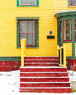 Red wooden steps covered in snow leading to bright yellow house with green trim and windows