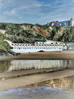 Beach huts lined up along Tolcarne Beach with cliff backdrop and reflections in wet sand