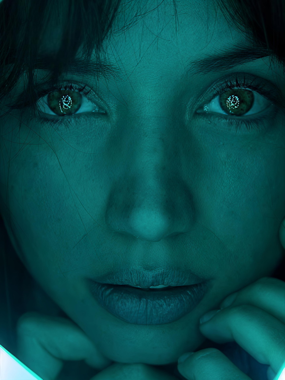 Close-up portrait of a young woman with intense eyes under teal-green lighting, hands beneath her chin