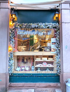 Decorated European shop front with olive branch garland, fairy lights, and wooden shelves displaying cheese and food items